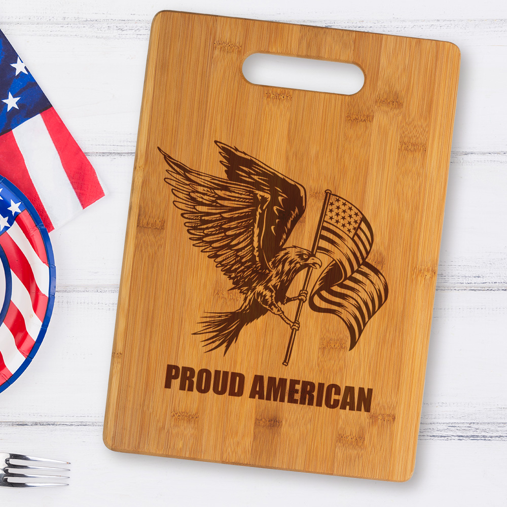 BCB2 bamboo cutting board engraved with an eagle holding an American flag with the text Proud American sitting on a table with patriotic plate and napkin