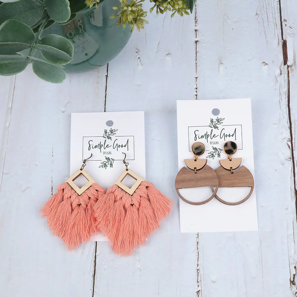 2 pairs of earrings in a cardholder sitting on a table. one is diamond shape with pink string fringe and the other is abstract shapes