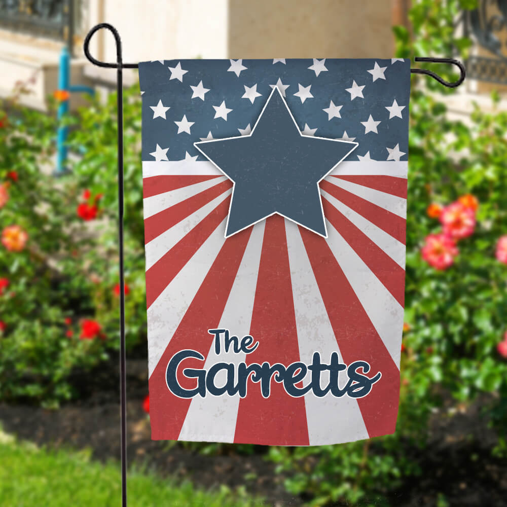 Patriotic garden flag hanging outside in front of flowers. Flag has last name "The Garretts" on it with a large blue star and American flag background