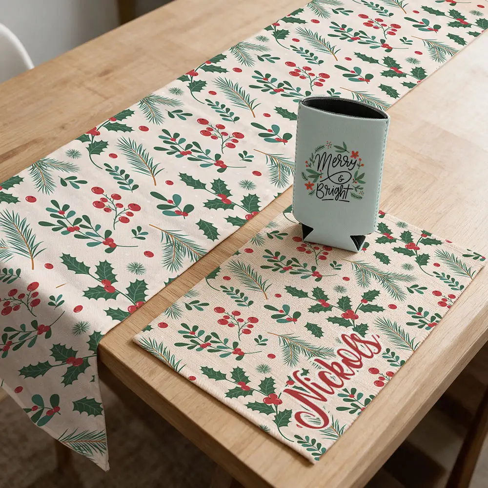 Christmas-themed table runner and placemat with evergreen branches, holly, and red berries, styled with a gray can cooler featuring “Merry & Bright” lettering and a personalized “Nickols” design on the placemat.