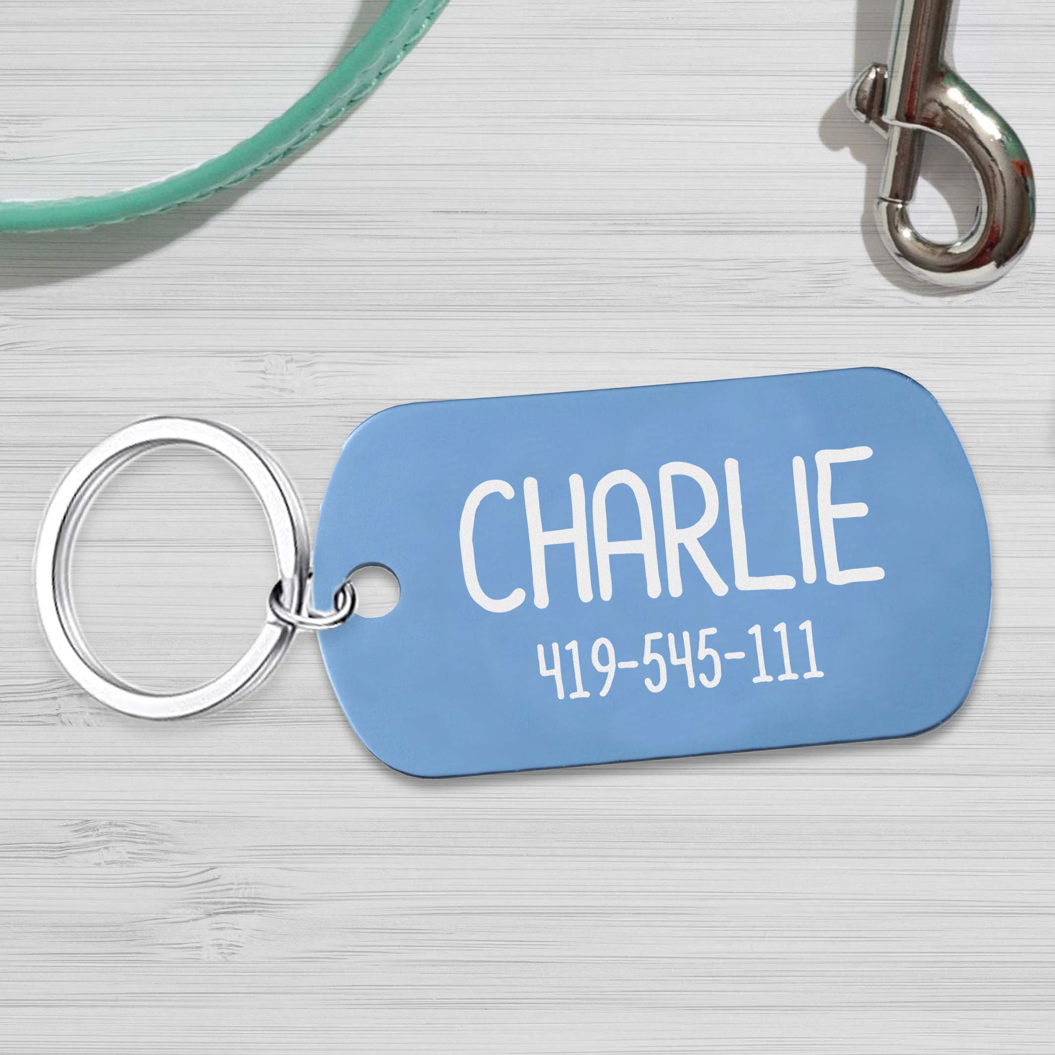 Light Blue anodized metal tag with the name Charlie engraved in it and a phone number. Tag is sitting on a table with a leash