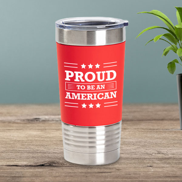 stainless steel tumbler sitting on a table with a red silicone sleeve on it and an engraving in white that says Proud to be an American
