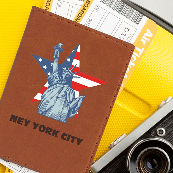 Saddle chestnut passport cover sitting on a yellow suit case with Statue of Liberty and American flag star printed on the cover with the engraving that says New York City
