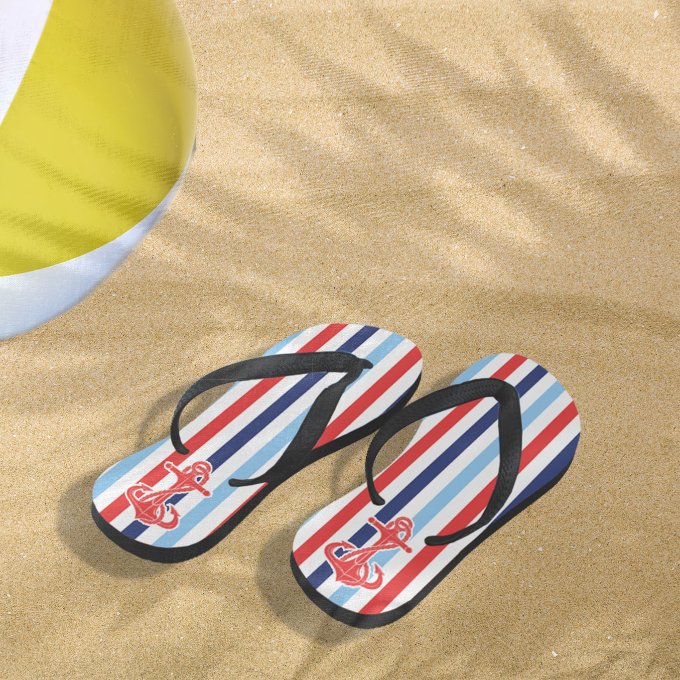 red white and blue anchor design printed on the top of flip flops. Flip flops sitting in sand on beach next to beachball