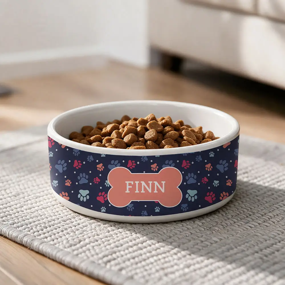 White ceramic pet bowl filled with dry food, featuring a navy blue exterior with colorful paw prints and a personalized bone-shaped nameplate reading “Finn.”