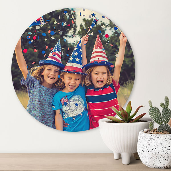 Circle photo panel with children celebrating the 4th of July printed on it hanging on white wall with plants sitting beside it