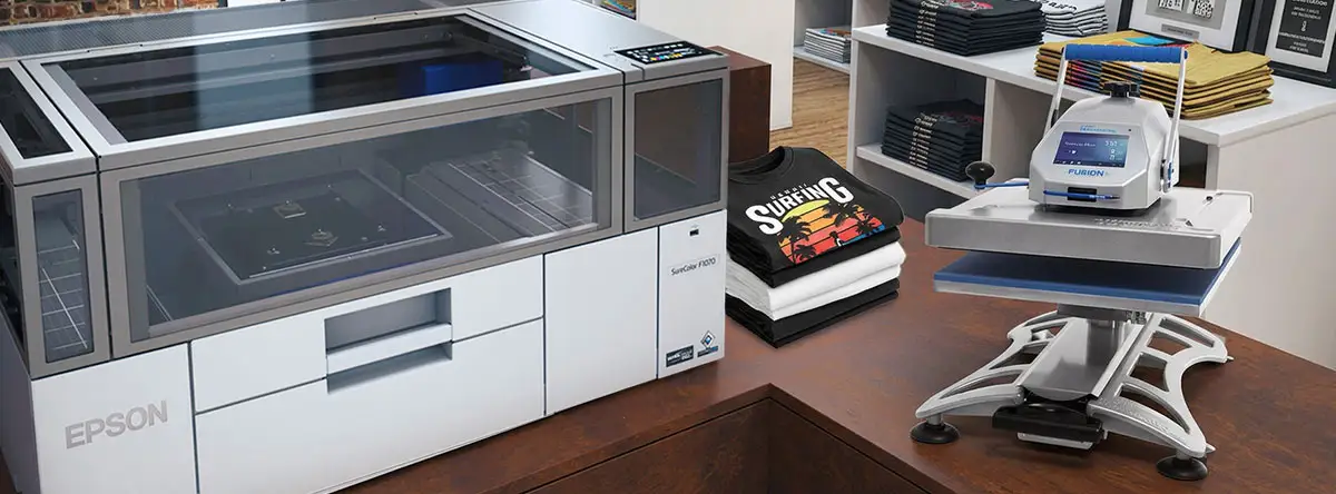 Epson F1070 DTG/DTF printer sitting on table with stack of T-Shirts and Fusion IQ heat press in a T-Shirt shop