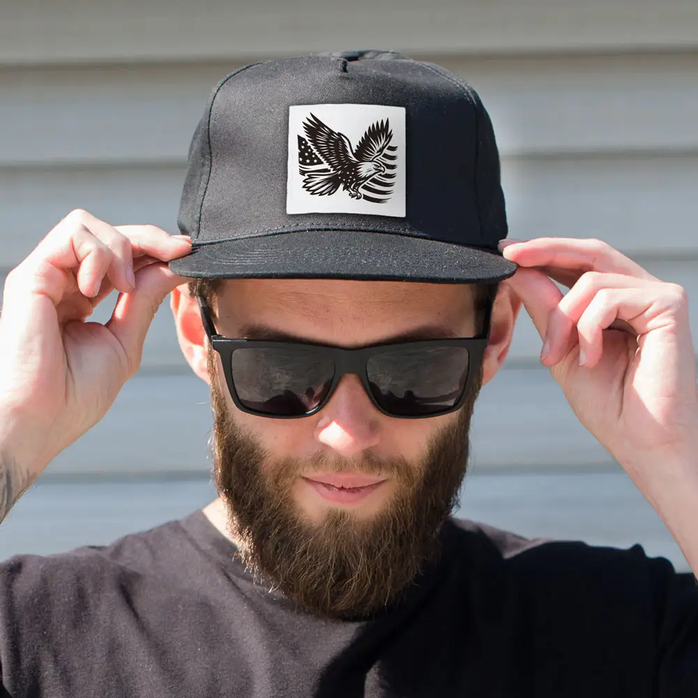 man wearing sunglasses holding his black cap that has a white patch on it with eagle and American flag engraved in black